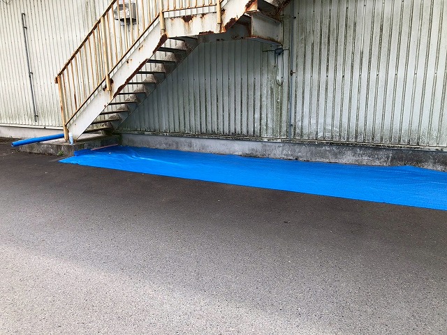 工場建物の鉄骨階段下の床面にブルーシートを敷いて養生している様子