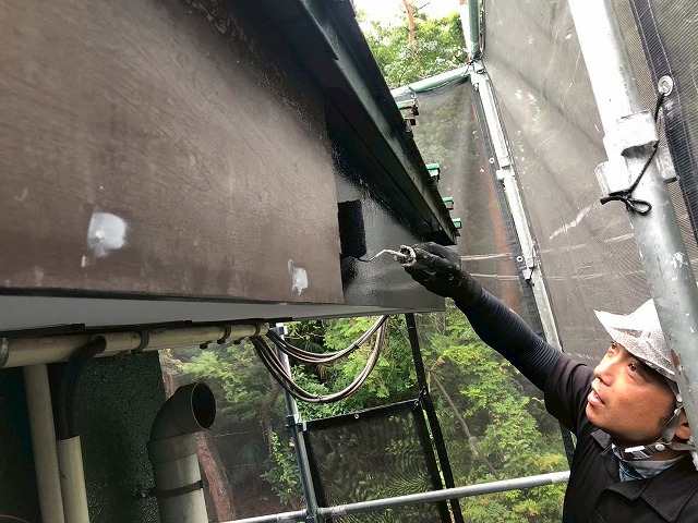足場上から破風板の側面に黒色塗料をローラーで塗り進めている様子