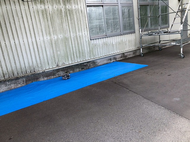 工場建物の壁際にブルーシートを敷いて養生し、移動式足場を設置している様子